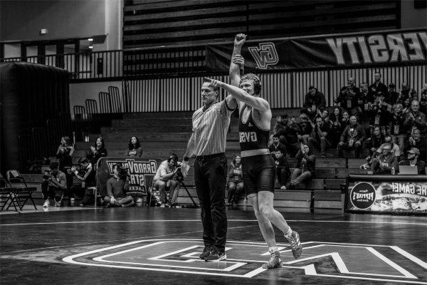 A referee holds up the arm of a person who just won a wrestling match. The person who won is smiling and pointing.