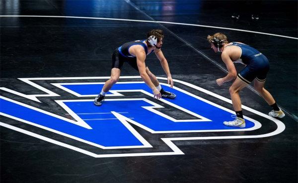 Two people bent in wrestling poses face each other on a mat with the letters GV.