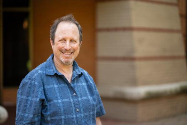 Joel Stillerman in a short sleeve blue shirt standing against a brick wall