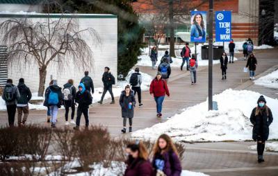 students walk on sidewalk, snow on grass areas; banner on lightpost reads Next is Now