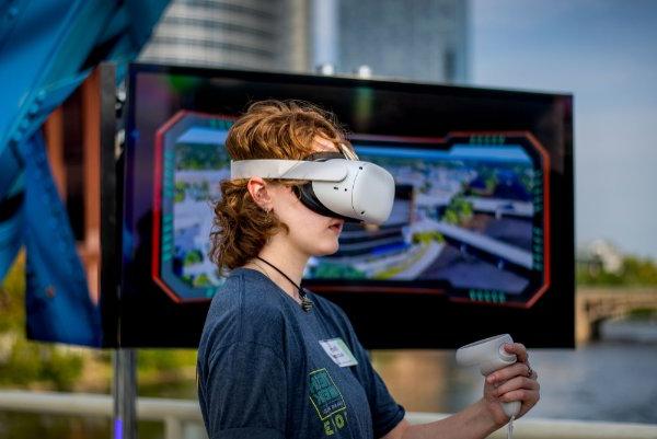 A person looks through a VR headset while standing before a screen.