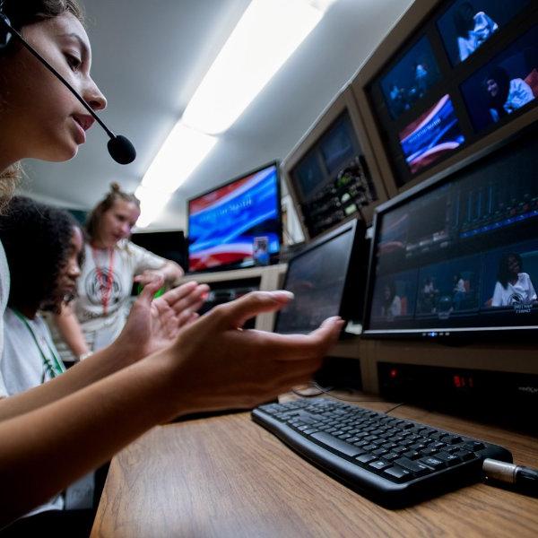 A person wearing a headset and looking at a computer screen claps. Two other people are in the background.