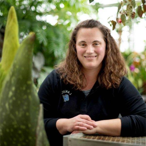 A person with arms folded on table smiles while standing next to a plant.