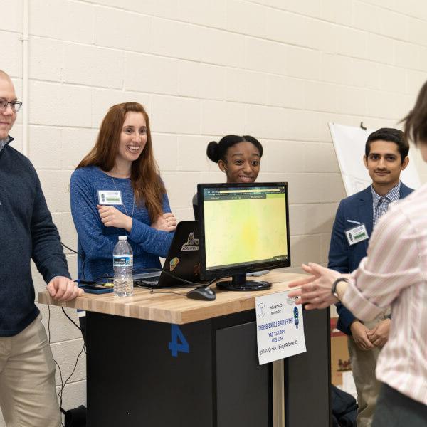 A group of students stand around a posterboard, describing work they did on an air quality monitoring program.