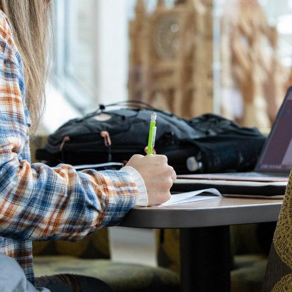 A person's hand holds a pen while the person writes on paper. A backpack and laptop are also on the table.