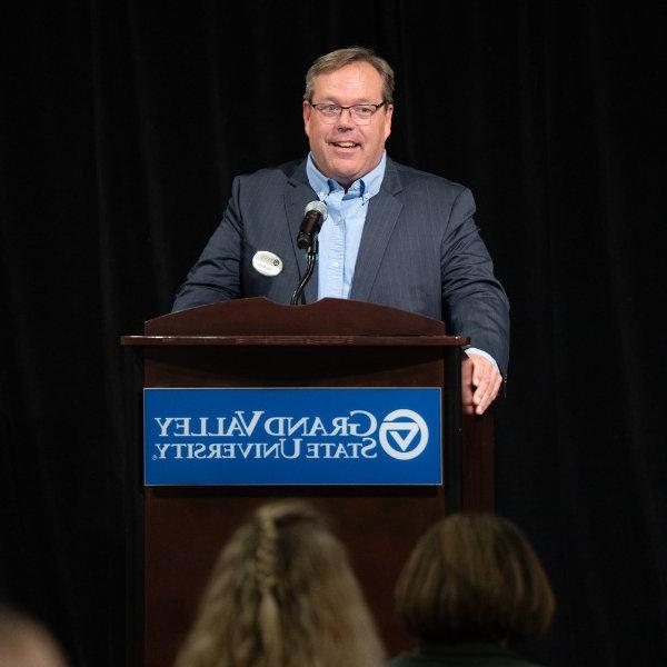 格雷格Warsen at podium in blue suit coat and light blue shirt with no tie; podium has GVSU wordmark on it